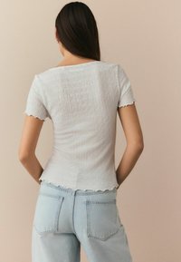 Top ajusté blanc texturé avec des manches courtes et des bords scallopés, associé à un jean taille haute bleu clair. Vue de dos.
