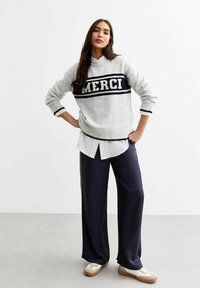 Ljusgrå stickad tröja med texten "MERCI", ihop med en vit randig skjorta, mörka vida byxor och krämfärgade sneakers med bruna sulor.