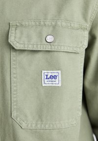 Green fabric shirt featuring a chest pocket with a silver snap button and a white label with blue text reading "Lee WORKWEAR."
