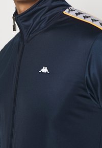  marinblå zip-up träningsjacka tillverkad av slät tyg, med en broderad vit logotyp och mönstrade axelaccenter i vitt och orange.