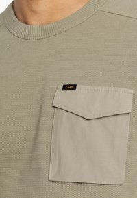 Khaki bluza z fakturowanej dzianiny, z kieszenią na piersi w jaśniejszym odcieniu. Czarna naszywka "Lee" na kieszeni. Okrągły dekolt.