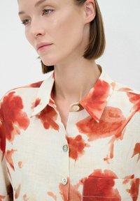 Bloemdruk shirt met rode en oranje tinten op een beige achtergrond, met een kraag, knopen en een borstzak.
