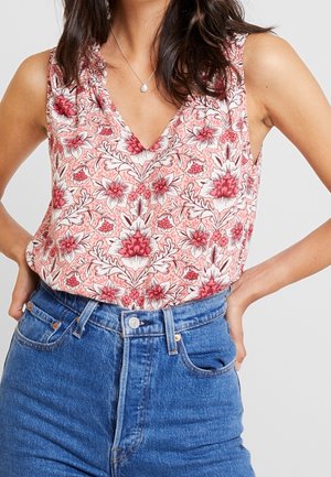 Mujer con blusa rosa sin mangas con estampado floral metida en vaqueros azules de talle alto, manos apoyadas en las caderas, mostrando la zona media.