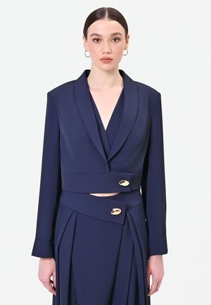Femme aux cheveux foncés portant un ensemble blazer et jupe bleu marine avec des boutons dorés et un col en V profond sur un fond clair uni.
