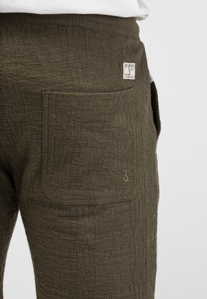 Nahaufnahme einer strukturierten dunkelgrünen Hosentasche mit kleinem Stoffetikett und gesticktem Logodetail auf weißem Hintergrund.