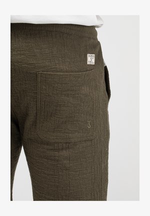 Nahaufnahme einer strukturierten dunkelgrünen Hosentasche mit kleinem Stoffetikett und gesticktem Logodetail auf weißem Hintergrund.