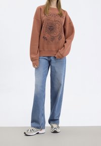 Hnědá mikina se grafickým designem slunce, spárovaná se světle modrými širokými džíny a vzorovanými teniskami. Jednoduchý, ležérní outfit.