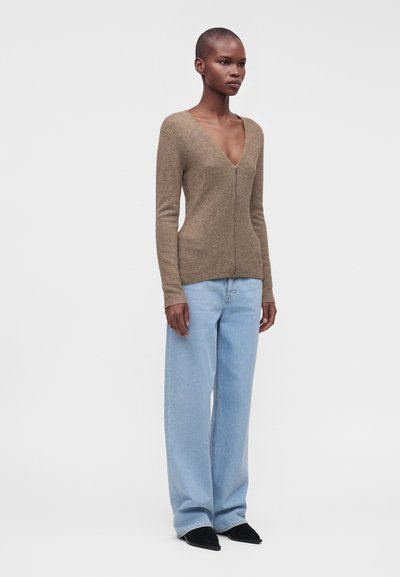 Femme debout portant un pull marron zippé avec un col en V profond, un jean bleu clair à jambes larges et des chaussures pointues noires sur fond blanc.