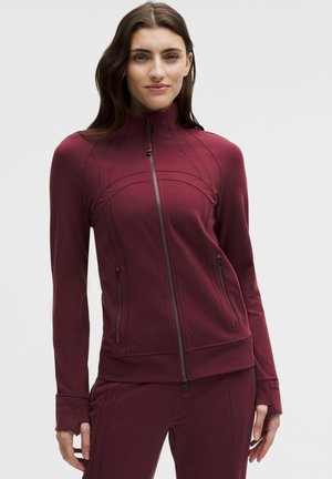 Kobieta w dopasowanej bordowej kurtce zapinanej na zamek i pasujących spodniach, stojąca z rękami swobodnie opuszczonymi wzdłuż ciała na jednolitym tle.