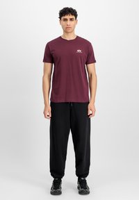 Burgundské bavlněné tričko s krátkým rukávem, s malým logem na hrudi. Spárováno s černými tepláky a černými sportovními botami.