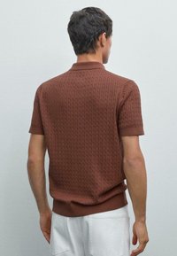 Brun kortärmad polo skjorta med texturerat kabelstickat mönster, ribbad krage och nederkant. Modellen har vita byxor och ses bakifrån.