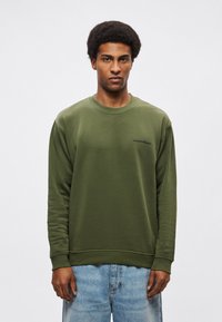 Sudadera verde oliva con mangas largas y cuello redondo, con un pequeño logo bordado en azul marino. Tela texturizada con puños de canalé.