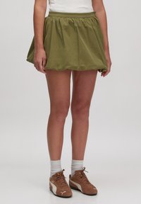 Groene, uitlopende mini-rok met een geplooide taille, gemaakt van lichte stof. Gecombineerd met bruine sportschoenen en witte crew-sokken.