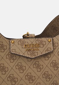 Brun mønstrede håndtaske med et hexagonalt mærke, guld hardware og en struktureret overflade. Mærkenavnet "GUESS" og "EST 1981" prægede.
