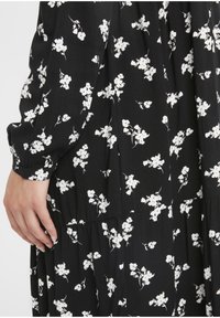 Schwarzes Blumenkleid mit weißen Blumenmustern, gerafften Volants, langen Ärmeln und einem weichen, strukturierten Stoff. Detailansicht des Ärmels und Saums.