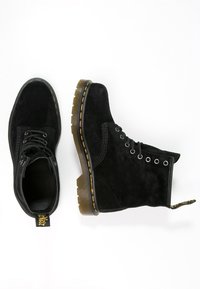 Botas de tobillo de ante negro con punta redonda, seis ojales y costuras amarillas. Presentan una suela marrón texturizada y una lengüeta con la marca.