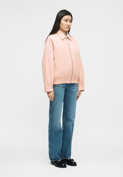 Γυναίκα με μακριά μαύρα μαλλιά στέκεται φορώντας ένα ανοιχτό ροζ μπουφάν με φερμουάρ, μπλε τζιν με ίσια μπατζάκια και μαύρα μοκασίνια, μπροστά σε ένα απλό φόντο.