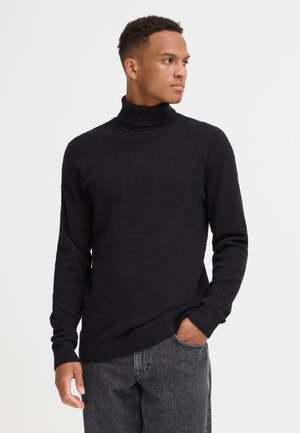 Maglione nero a collo alto in maglia con motivo testurizzato, polsini e orlo a coste, abbinato a jeans grigi. Il modello si trova in piedi con la mano in tasca.