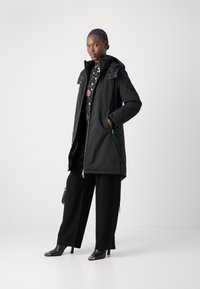 Black parka with a hood, zipper closure, side pockets, and adjustable waist. Paired with wide-leg black pants and black heeled shoes.