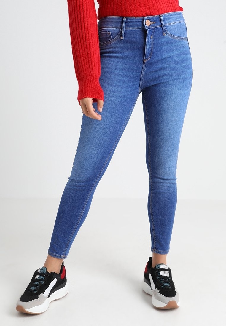 Höga blå jeans med slim fit, tillverkade av denim; kombinerat med en röd ribbad tröja och svarta sportiga sneakers.