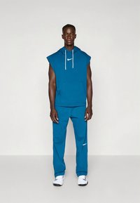 Turkizna Nike športna obleka s topom brez rokavov z kapuco in sprednjim žepom ter ujemajočimi hlačami. Obe imata subtilne logotipne poudarke.