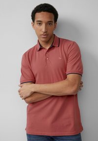 Roza polo majica s strukturirano ovratnikom, ki ima mornarsko modro akcentsko barvo, kratkimi rokavi in dvema gumboma. Tkanina izgleda gladko in rahlo elastično.