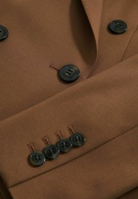 Ruskea räätälinpuku, jossa on sileä kangas, mustat napit, joissa on embossed-brändinimi, sekä neljä koristeellista napin yksityiskohtaa.