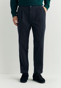 Donkerblauwe broek met een ruime pasvorm, voorzien van een elastische tailleband en voorplooien, gecombineerd met zwarte schoenen met zachte zolen.