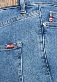 Detailní záběr zadní kapsy světle modrých džínů z denimu s červenými štítky značky "Big Star" a kovovým nýtem.