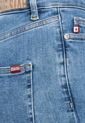 Valge taustaga hele sinise teksade tagatasku lähivaade, kus on punased "Big Star" kaubamärgiga sildid ja metallist nikerdus detail.