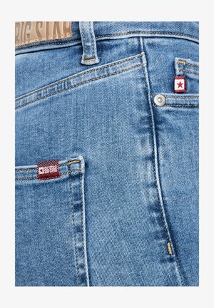 Valge taustaga hele sinise teksade tagatasku lähivaade, kus on punased "Big Star" kaubamärgiga sildid ja metallist nikerdus detail.