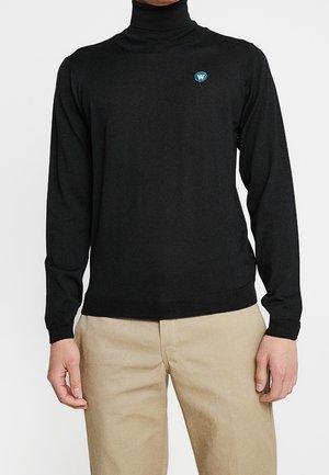 Mand iført en sort rullekravebluse med et blåt logo på brystet og beige bukser, stående mod en hvid baggrund.