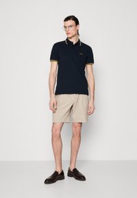 Polo shirt marine avec col rayé jaune, logo sur la poitrine, associé à un short beige et des mocassins marron foncé. Le mannequin se tient devant un fond uni.