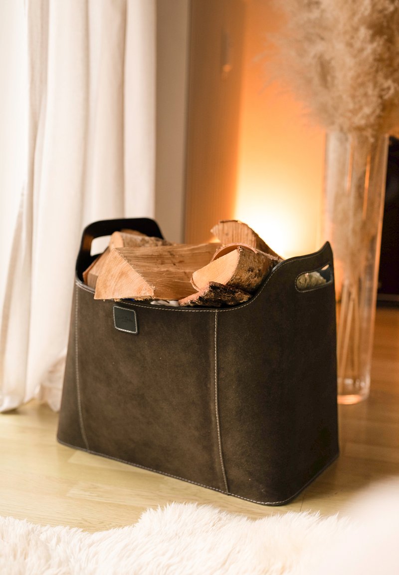 Ciemnobrązowy zamszowy kosz na drewno do kominka z zaokrąglonymi krawędziami i bocznymi uchwytami, wypełniony ułożonym drewnem opałowym. Widoczny charakterystyczny detal w postaci szwów.