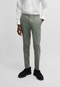 Pantalon ajusté vert clair avec un motif à carreaux subtil, fabriqué dans un tissu lisse. Porté avec une chemise blanche à boutons et des chaussures noires.