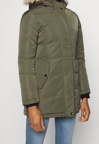 Olivgrön vadderad jacka med en huva av fuskpäls, dragkedja och knäppning, två sidofickor och elastiska ärmslut. Slät textur.