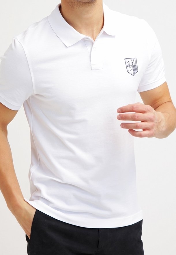 Biała koszulka polo z kołnierzykiem, krótkimi rękawami i granatowym emblematem po lewej stronie klatki piersiowej. Materiał jest gładki i lekki, a wzór klasyczny.
