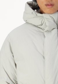 Chaqueta acolchada gris claro con un alto cuello, tejido texturizado y un diseño minimalista con cordón ajustable para un ajuste personalizable.