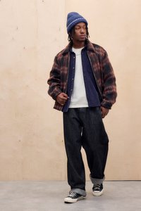 Rudet brun og sort jakke, marineblå skjorte, hvid t-shirt, løse mørke jeans, blå strikket hue og sorte sneakers med hvid sål.