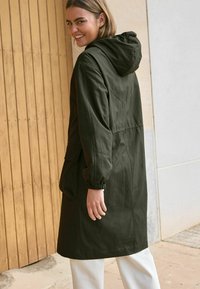 Giacca verde scuro con cappuccio, polsini elasticizzati, vita cinchata e due grandi tasche frontali. Tessuto liscio con dettagli di cucitura minimi.