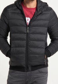 Schwarze Steppjacke mit horizontalen Nähten, durchgehendem Reißverschluss vorne und Kapuze. Verfügt über elastische Bündchen und Saum, getragen über einem roten Sweatshirt.