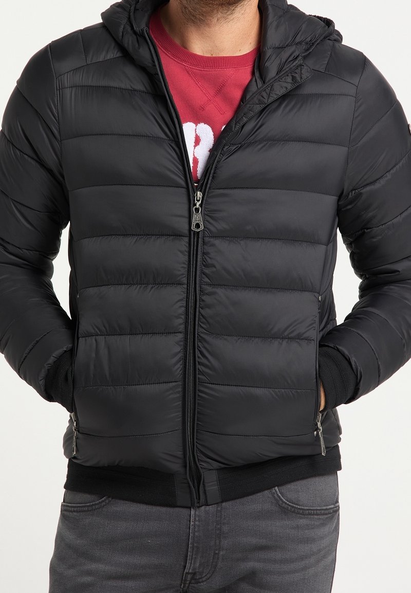 Schwarze Steppjacke mit horizontalen Nähten, durchgehendem Reißverschluss vorne und Kapuze. Verfügt über elastische Bündchen und Saum, getragen über einem roten Sweatshirt.