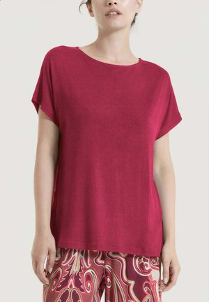 Donna che indossa una maglietta bordeaux a maniche corte ampia e pantaloni fantasia rossi, beige e bordeaux, in piedi davanti a uno sfondo uniforme.