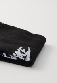 Μαύρη πλεκτή κάλτσα με χοντρή υφή, που έχει λευκή κεντημένη λογότυπο κοντά στην άκρη. Η κάλτσα είναι διπλωμένη σε ορθογώνιο σχήμα.