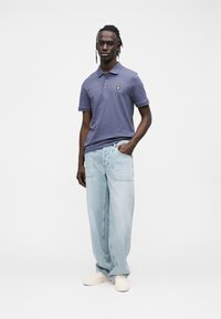 Polo azul escuro com mangas curtas e um logótipo amarelo; combinado com jeans largos azul claro e sapatilhas bege.