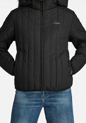 Personne portant une veste matelassée noire avec une fermeture éclair à l'avant et un jean bleu, mains dans les poches de la veste, visage non visible.