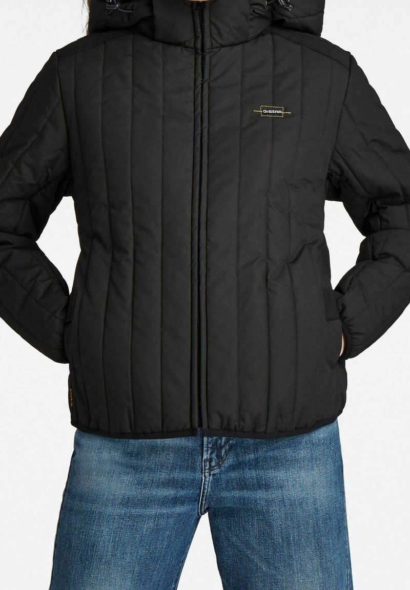 Personne portant une veste matelassée noire avec une fermeture éclair à l'avant et un jean bleu, mains dans les poches de la veste, visage non visible.