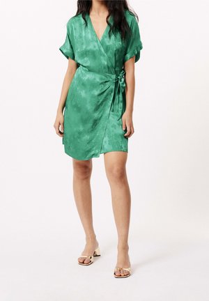 Femme portant une robe courte verte portefeuille avec un nœud sur le côté et des manches courtes, assortie à des sandales beige à talons bloc et bouts ouverts.
