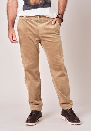 Man draagt beige corduroy broek, donkerbruine veterlaarzen met witte zolen en een wit T-shirt met een grafische print.