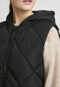 Czarna pikowana kamizelka z kapturem i zapięciem na zamek, zestawiona po prawej stronie z kremowym dzianinowym swetrem. Widoczne są teksturowane tkaniny.